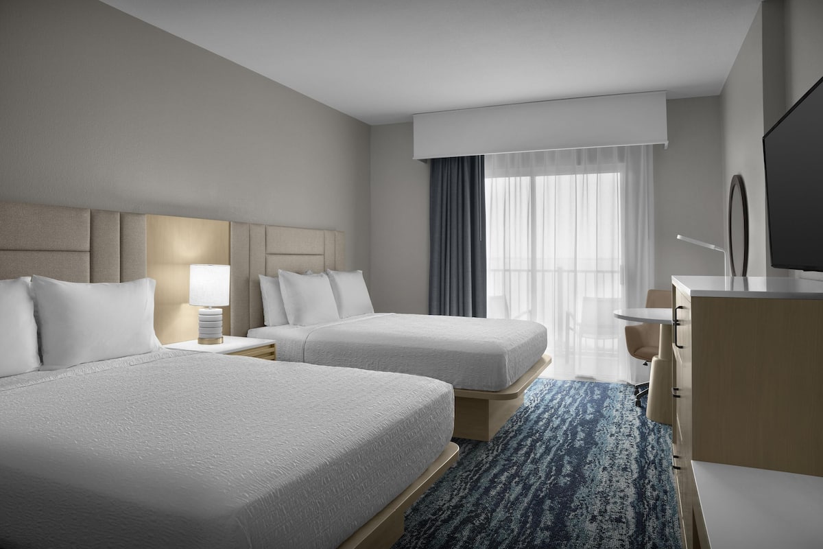 A hotel room with two beds, a desk, a TV, and a large window with curtains.