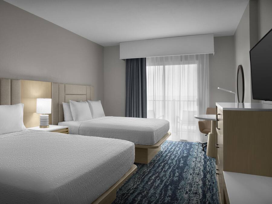 A hotel room with two beds, a desk, a TV, and a large window with curtains.