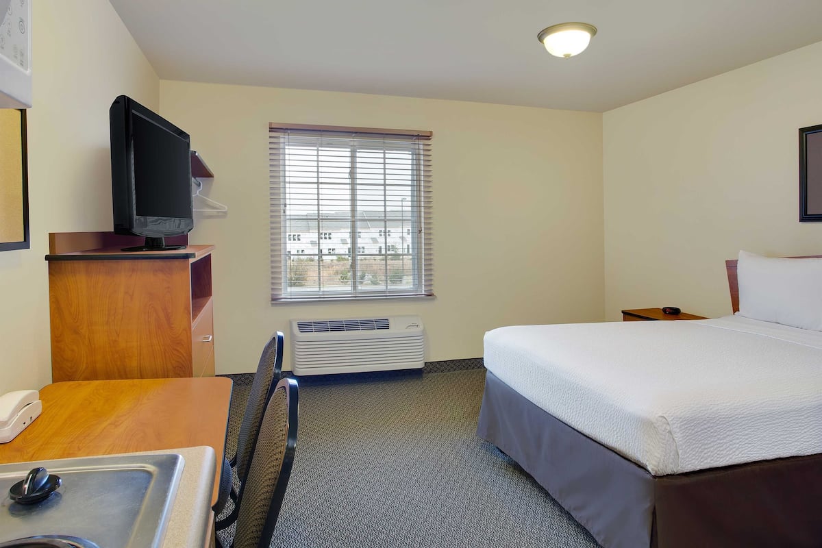 A hotel room with a bed, a desk with a telephone, a television, a window with blinds, and a ceiling light.