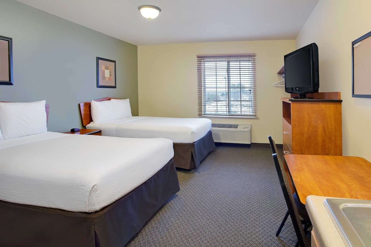 A hotel room with two beds, a television on a wooden stand, a desk, and a window with blinds.