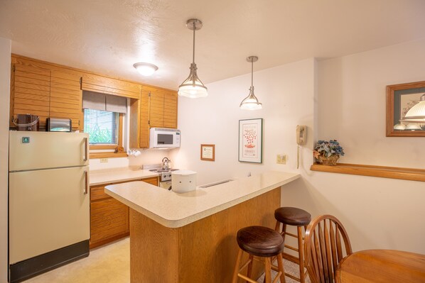 Una cocina con armarios de madera, un refrigerador blanco, un microondas, un lavabo y dos taburetes de bar.