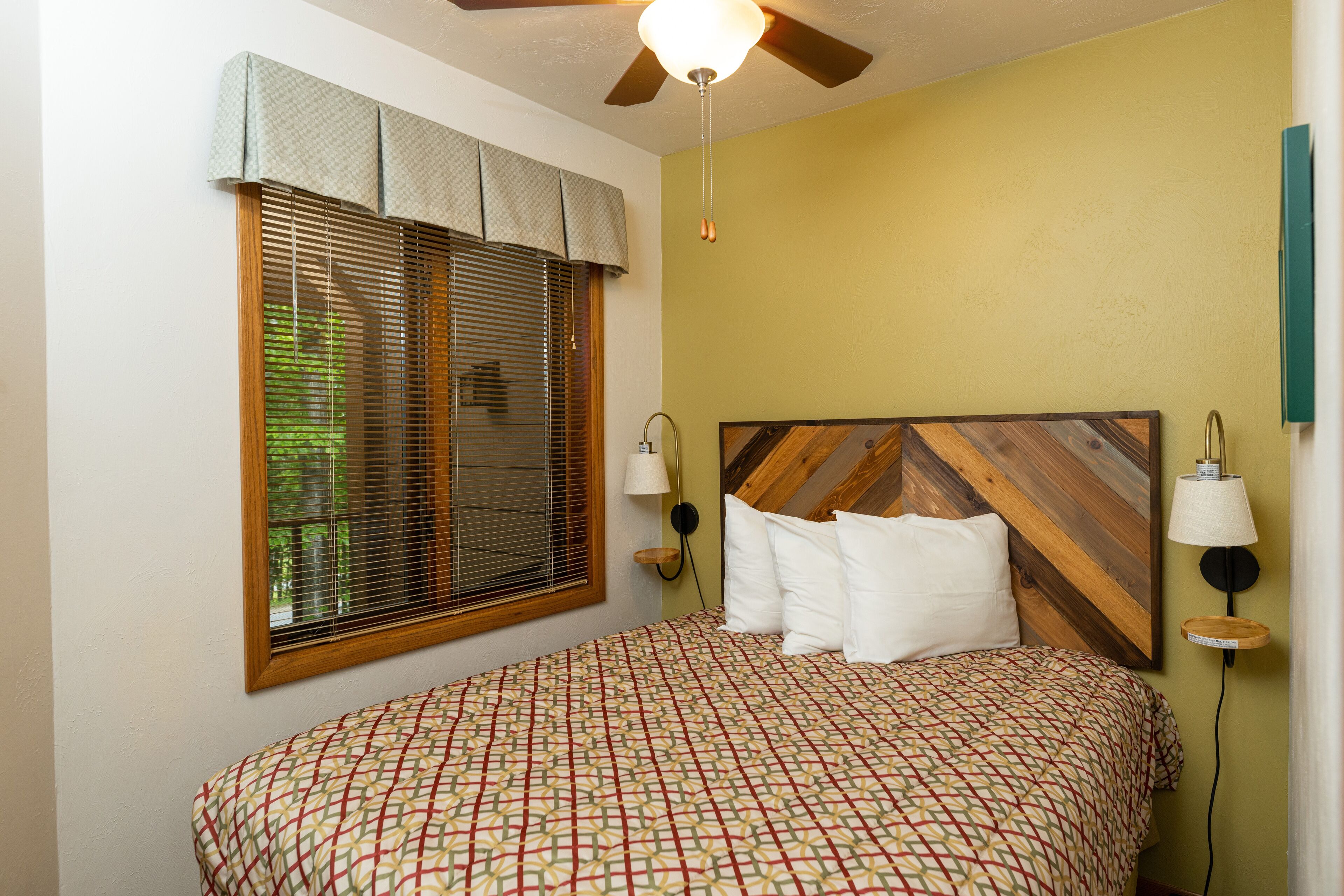 A bedroom with a bed, a ceiling fan, a window with blinds, and a wall-mounted lamp.