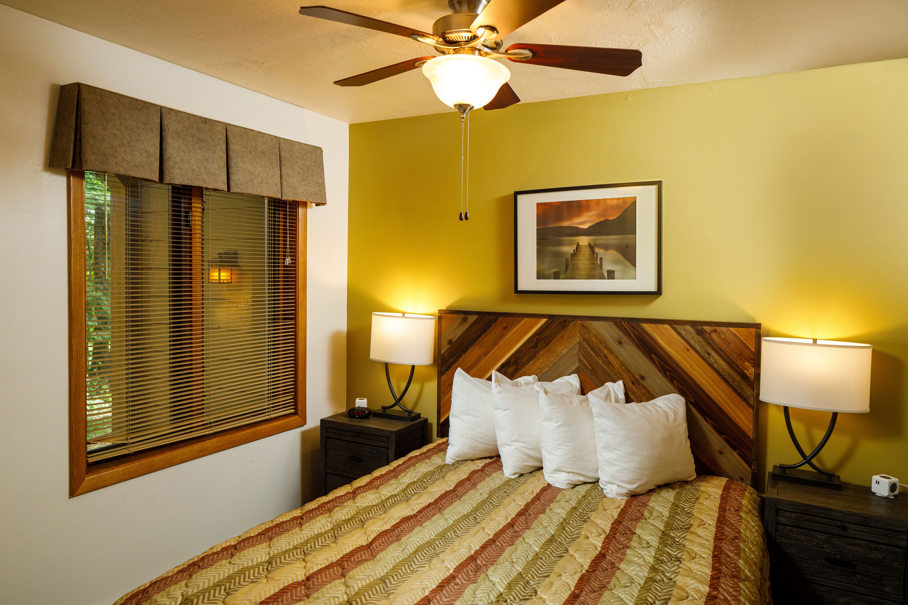 A bedroom with a bed, two bedside tables, a ceiling fan, a window with blinds, and a framed picture on the wall.