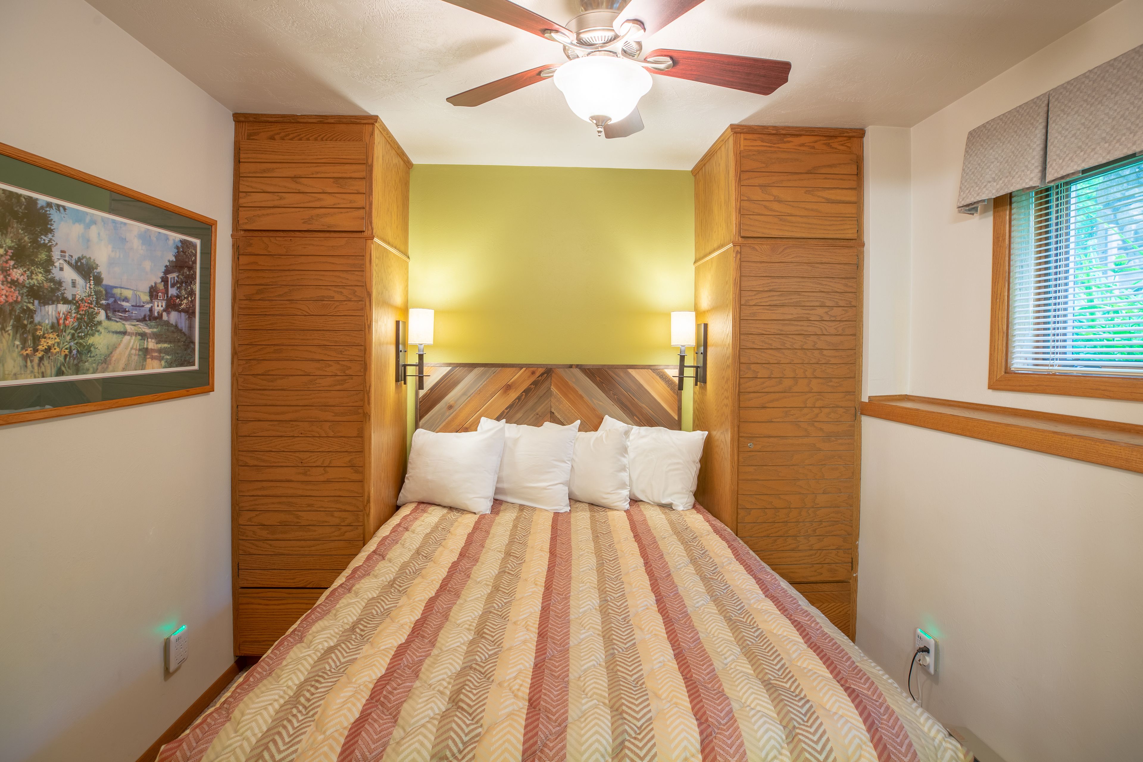 Una camera da letto con una testiera in legno, un letto con copripiumino a righe, luci a parete e una ventola a soffitto.