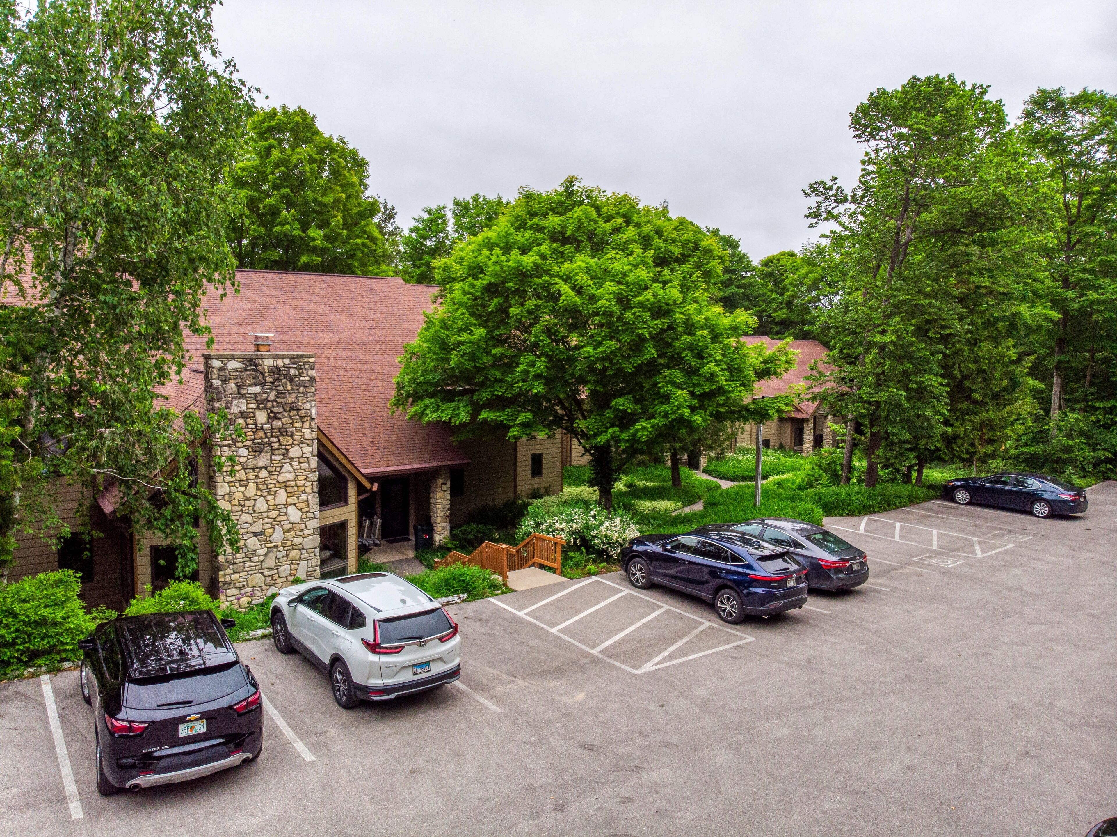 A parking lot with several cars, a stone building with a red roof, and lush green trees.