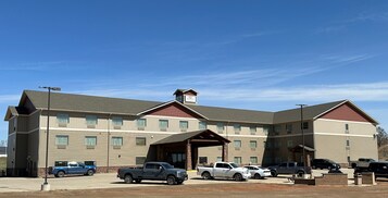 A multi-story hotel with a central entrance, multiple parking spaces, and a clear blue sky.
