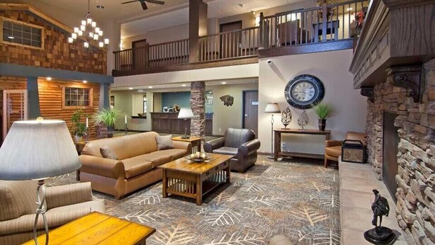 A spacious lobby with a high ceiling, a central seating area with sofas and a coffee table, a staircase leading to an upper level, and a stone fireplace. - The Watford (Watford City)