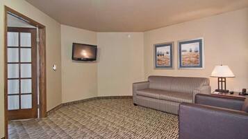 A hotel room with a patterned carpet, a sofa, a chair, a TV mounted on the wall, and two framed pictures on the wall.