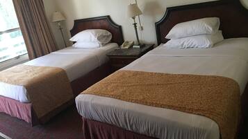 A hotel room with two beds, each with a wooden headboard, white linens, and a brown throw blanket. There is a nightstand with a lamp and a telephone between the beds.