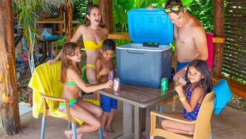 A group of people gathered around a table with a cooler, in a tropical setting with wooden furniture and greenery.