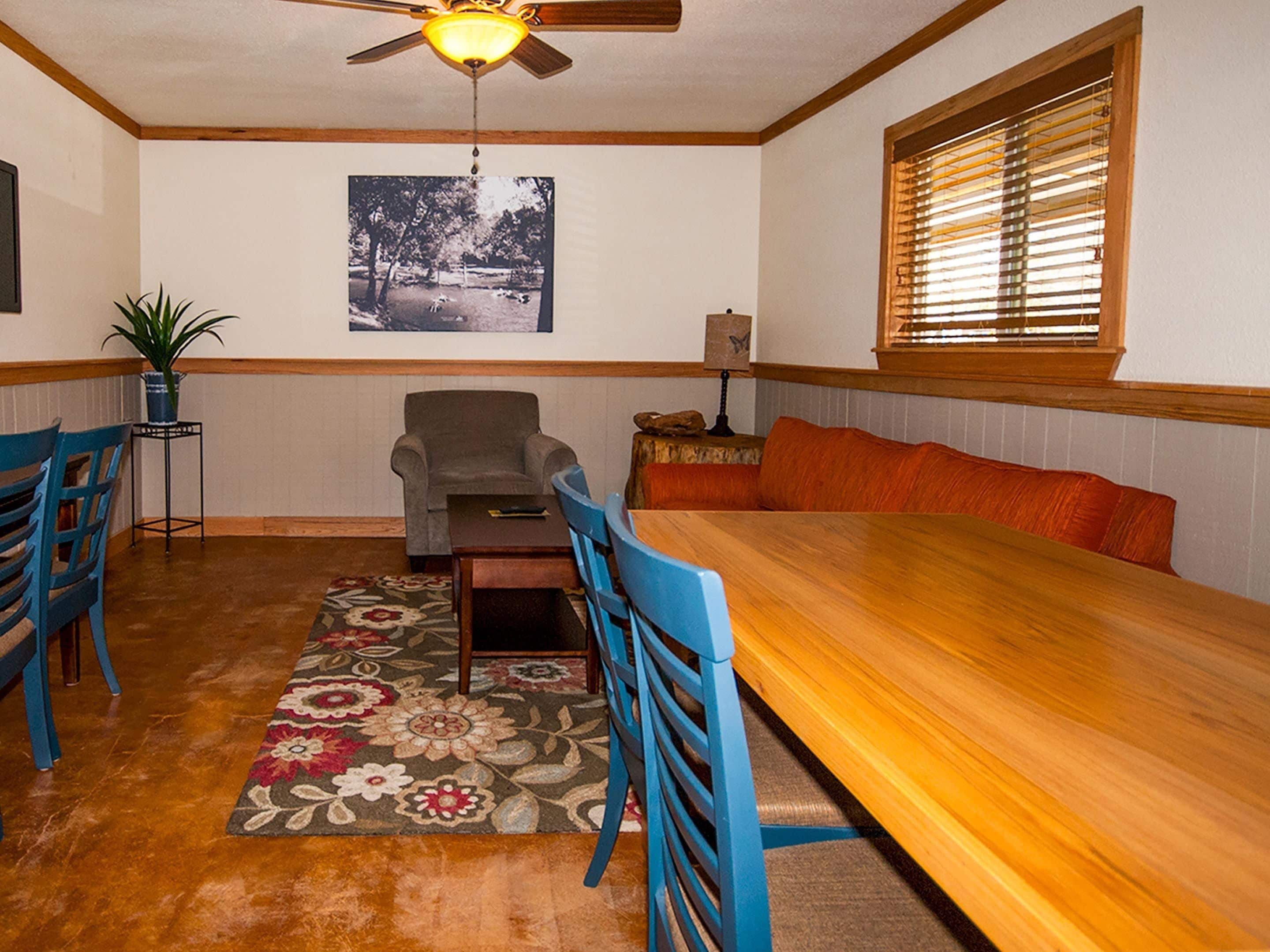 A dining area with a wooden table, blue chairs, a floral rug, a ceiling fan, a framed picture on the wall, and a window with blinds.