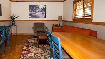 Un comedor con una mesa de madera, sillas azules, un tapete floral, un ventilador de techo, un cuadro en la pared y una ventana con persianas.