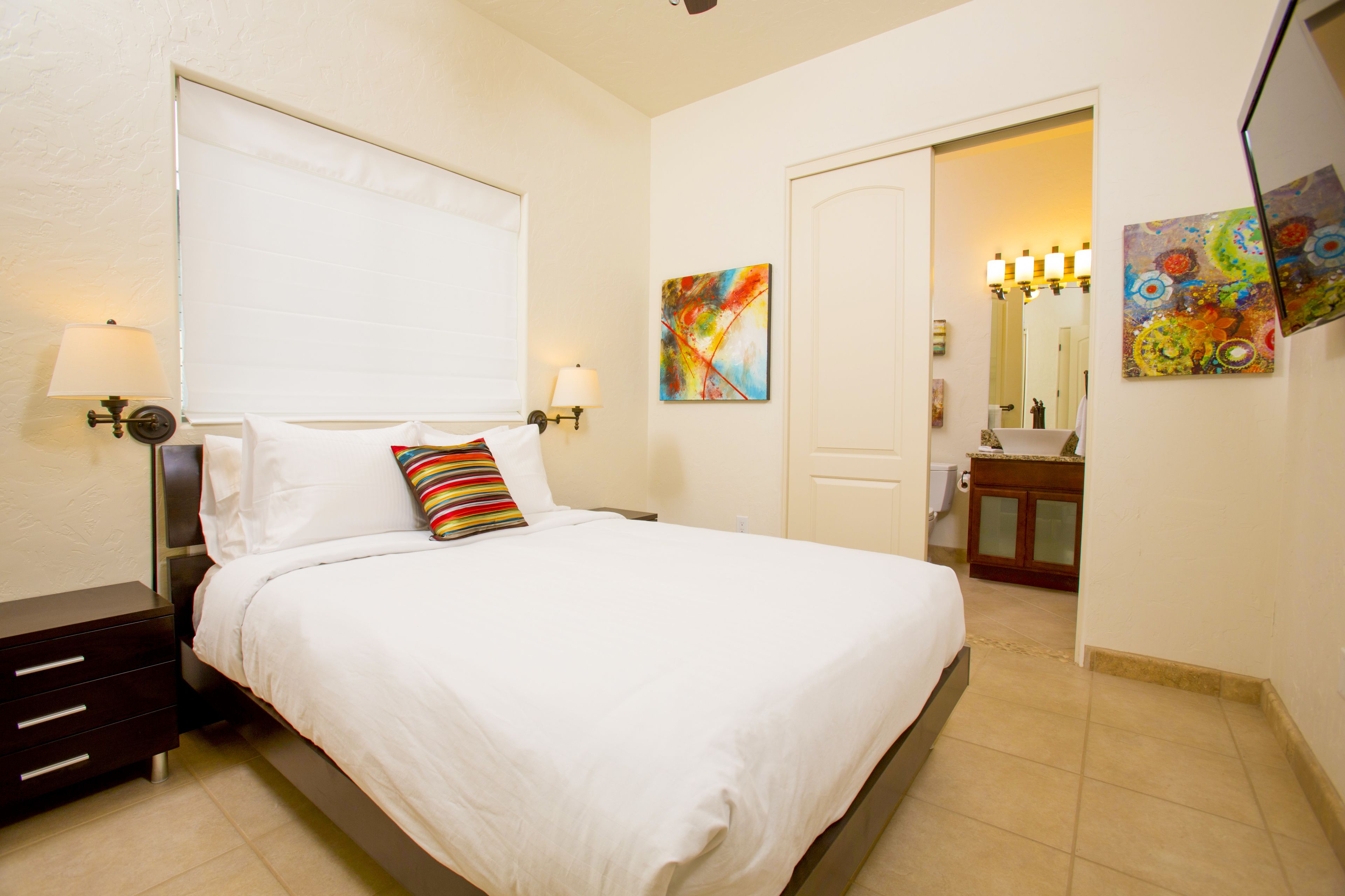 Habitación de hotel con cama, mesitas de noche, lámpara, un cuadro, y baño con espejo y lavamanos.