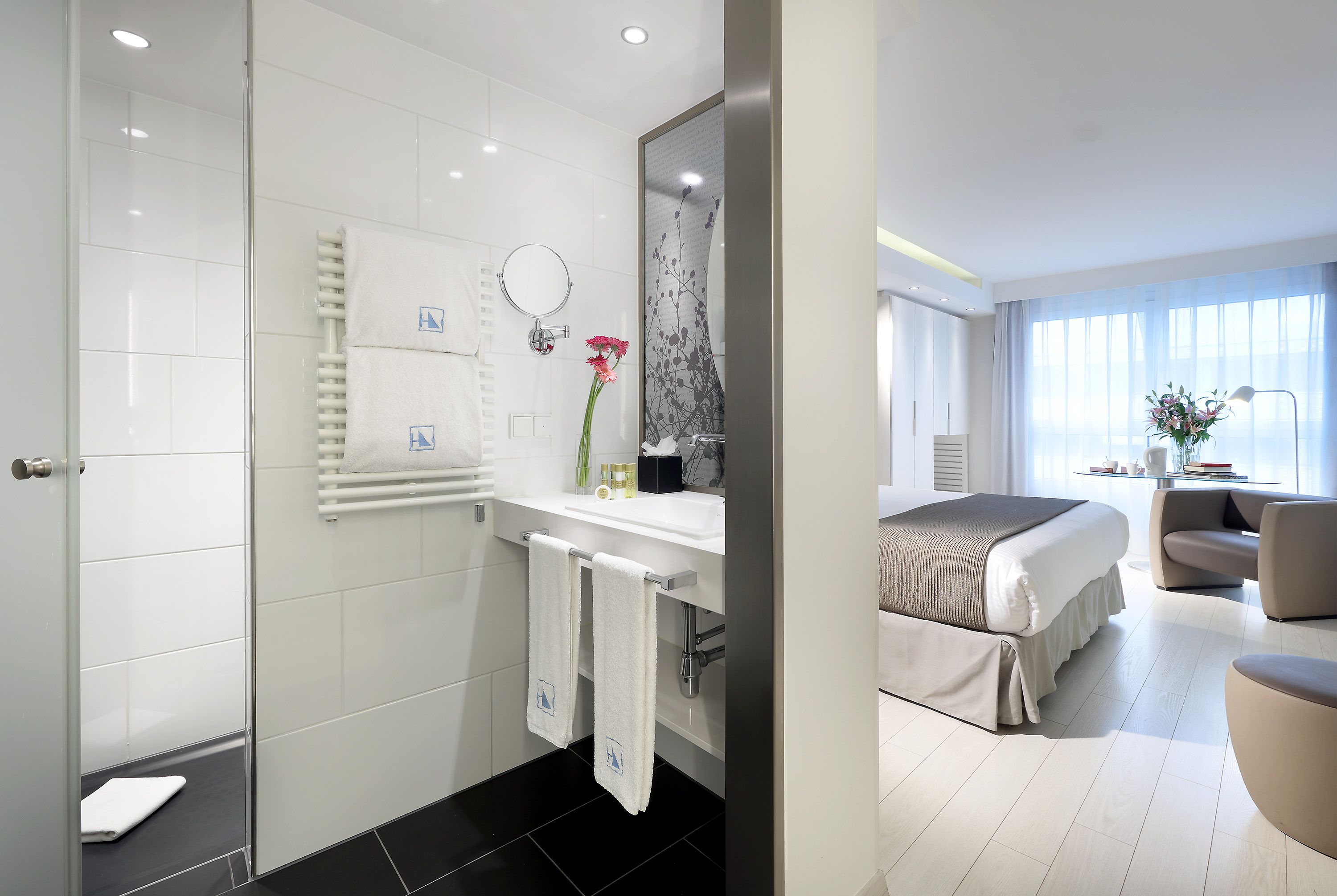 Ein modernes Hotelzimmer mit Dusche, einem Bett, einer Sitzecke und einem großen Fenster.