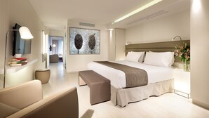 A modern hotel room with a large bed, a bedside table with a vase of flowers, a seating area with a bench, and a flat-screen TV mounted on the wall.