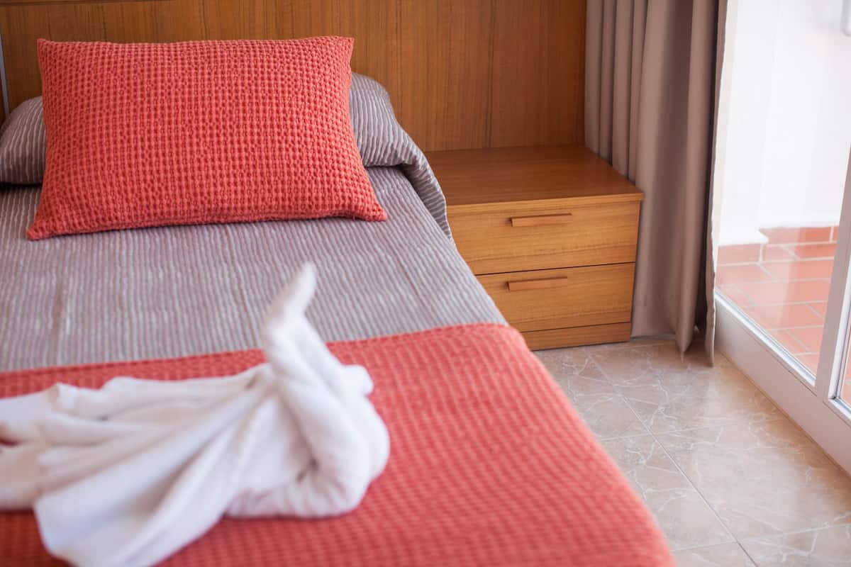 A neatly made bed with a red and grey blanket, a wooden nightstand, and a window with curtains.