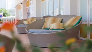 A wicker lounge chair with cushions, positioned on a tiled patio with a view of a building with large windows. - Hostal Los Pinos (Santa Eulalia del Rio)