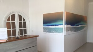 A reception desk with a wooden top and grey drawers, a white arched window, and two framed paintings of a coastal scene. - Hostal Los Pinos (Santa Eulalia del Rio)