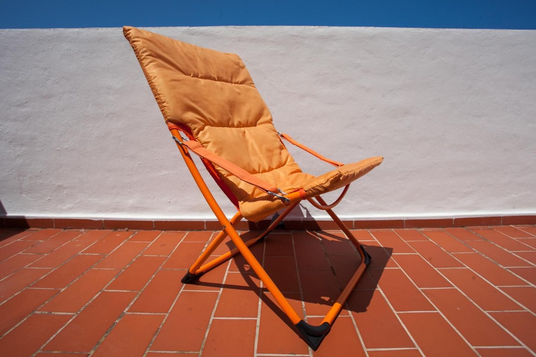 A single orange folding chair with a cushion, positioned on a red brick floor against a white wall.
