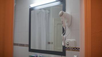 A bathroom with a hairdryer mounted on the wall, a mirror, and a shelf with toiletries.