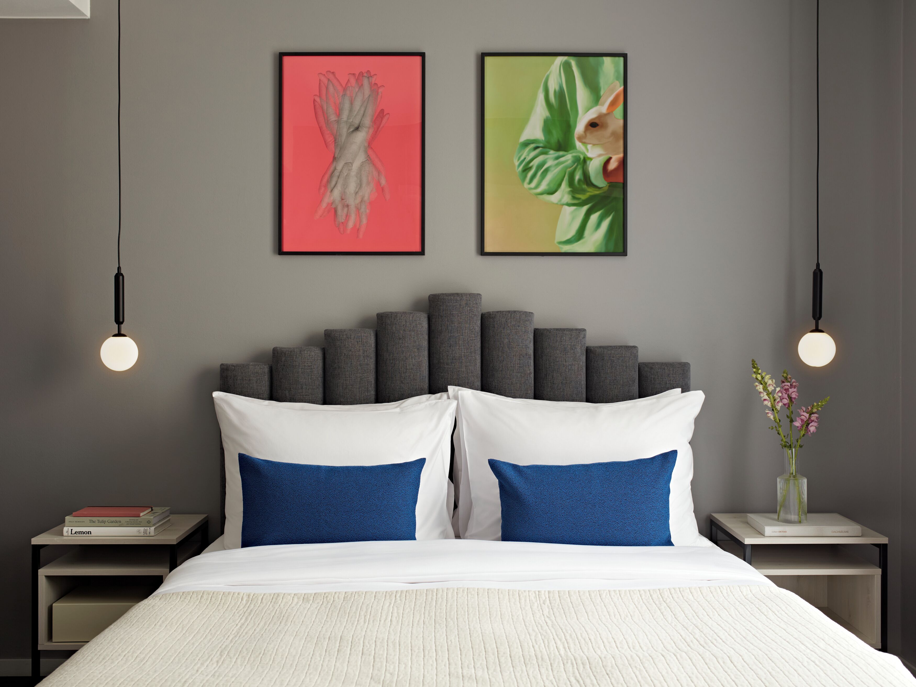 A modern bedroom with a grey headboard, white and blue pillows, a white bedspread, and two framed artworks on the wall.