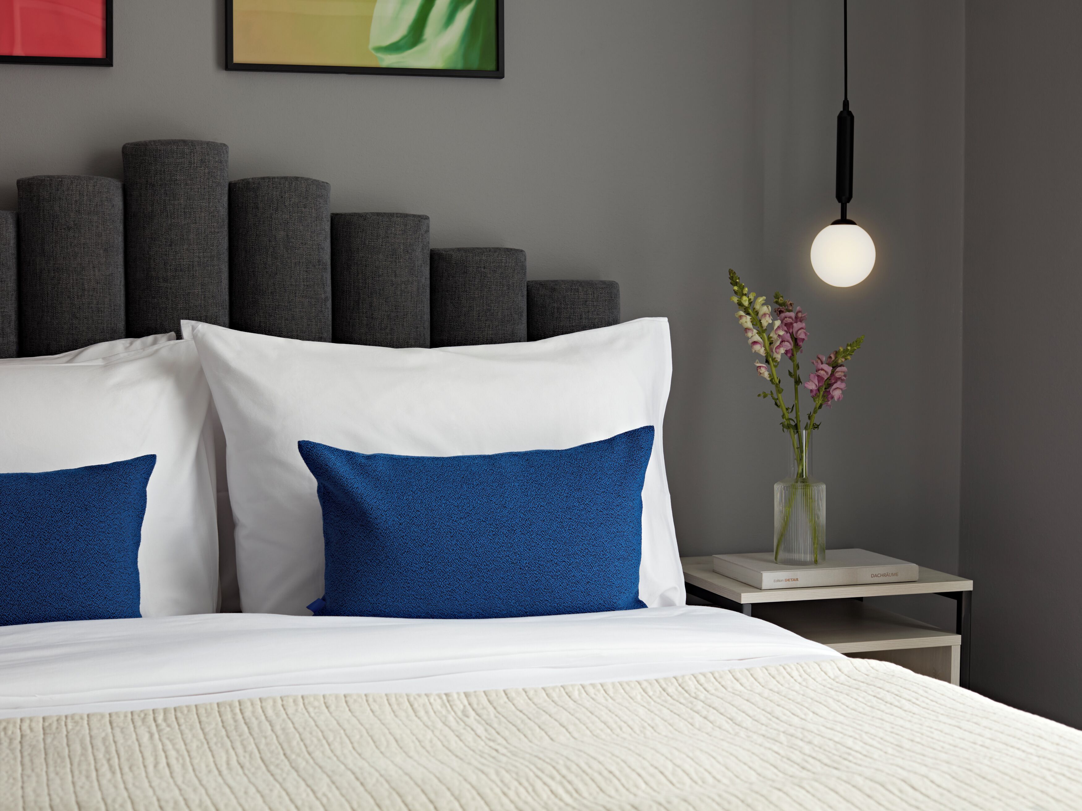 Ein modernes Schlafzimmer mit Bett, Kissen, einem Nachttisch, einer Vase mit Blumen und einer Wandleuchte.