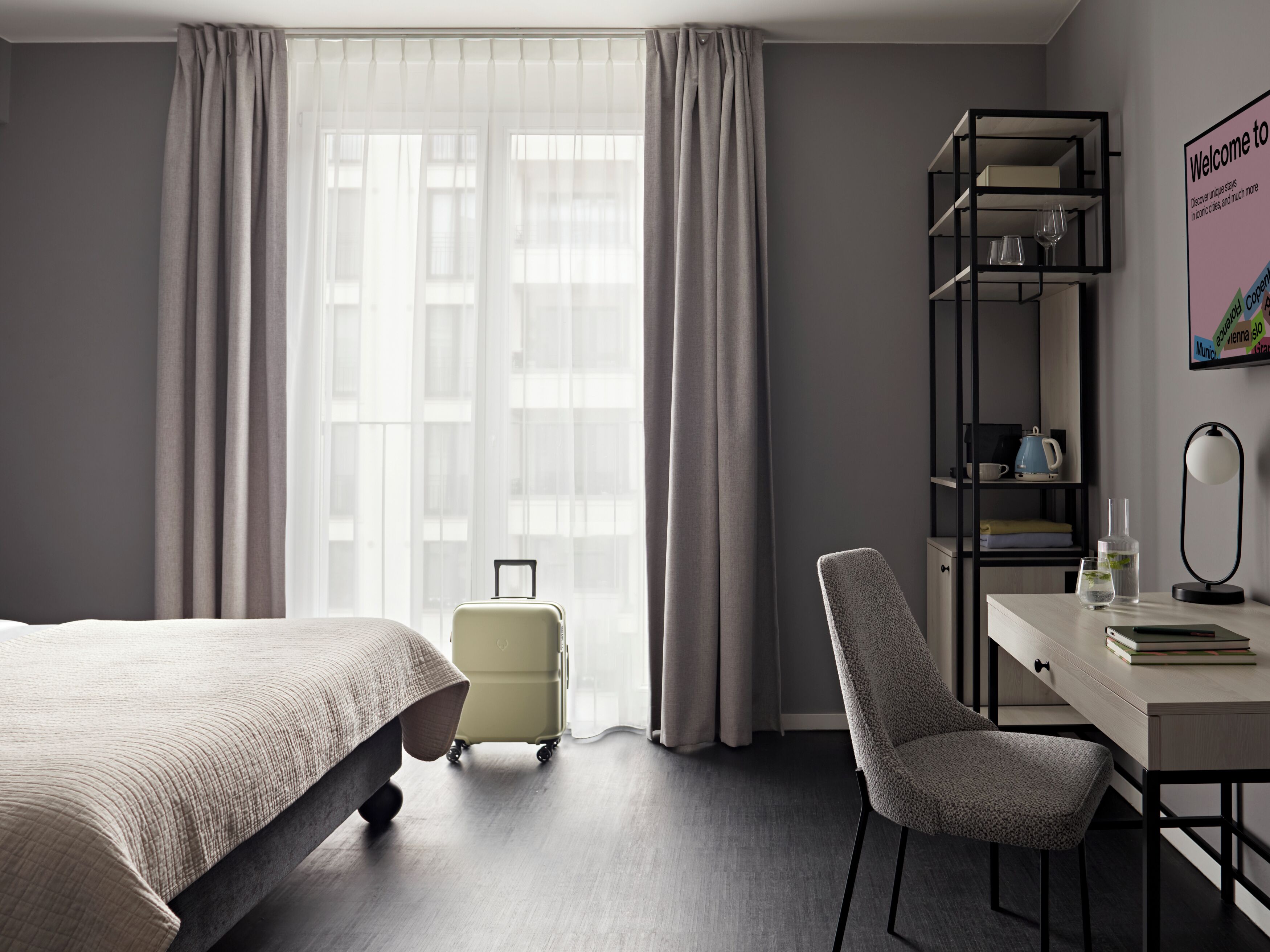A hotel room with a bed, a desk, a chair, a suitcase, and a bookshelf.