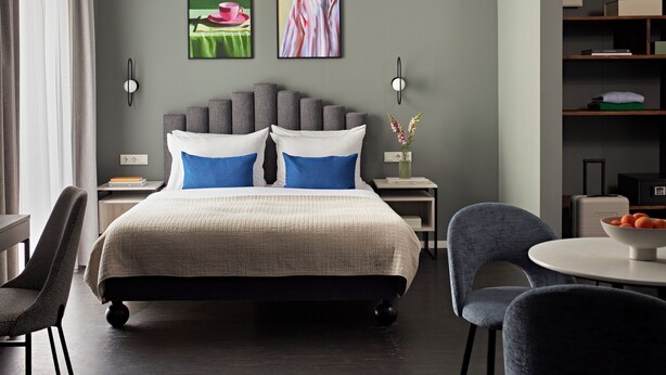 A modern hotel room with a large bed, two side tables, a small round table, and a chair. There are two wall-mounted light fixtures and a vase with flowers on the side table. - Numa Berlin Boxer (Berlin)