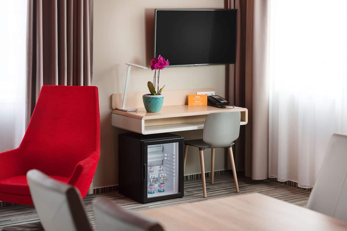 A hotel room with a desk, a red chair, a small refrigerator, a potted orchid, a flat-screen TV, and a city view.
