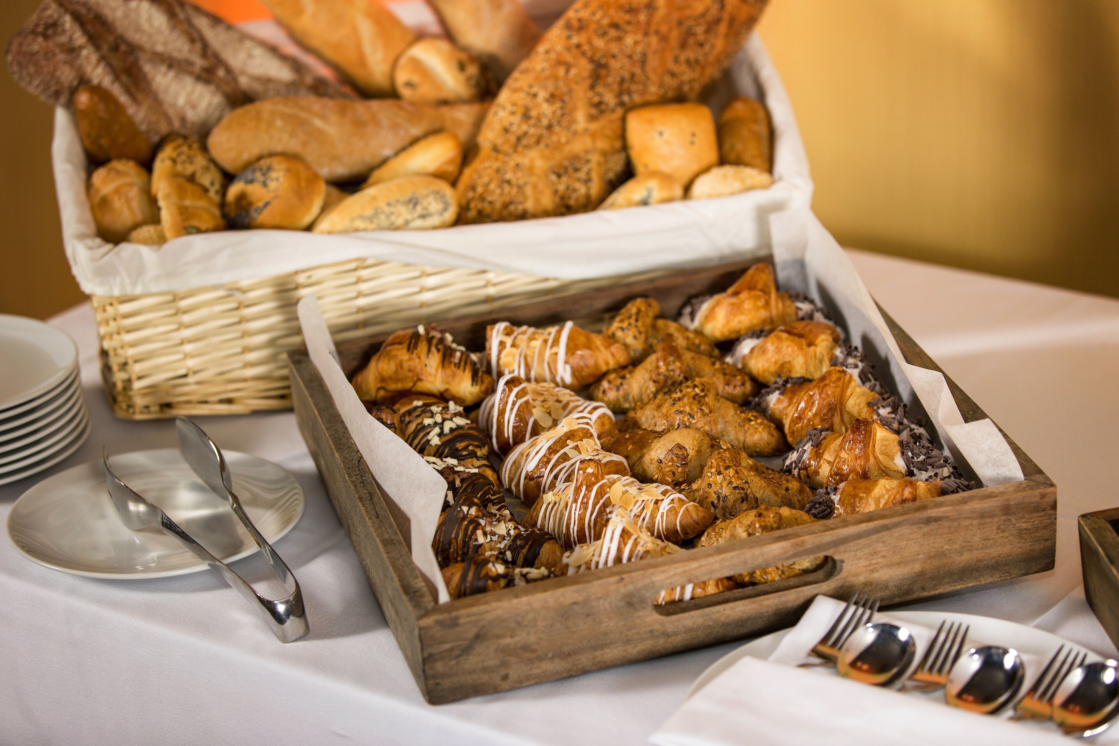 A wooden tray filled with various pastries and bread, accompanied by silverware and plates on a table.