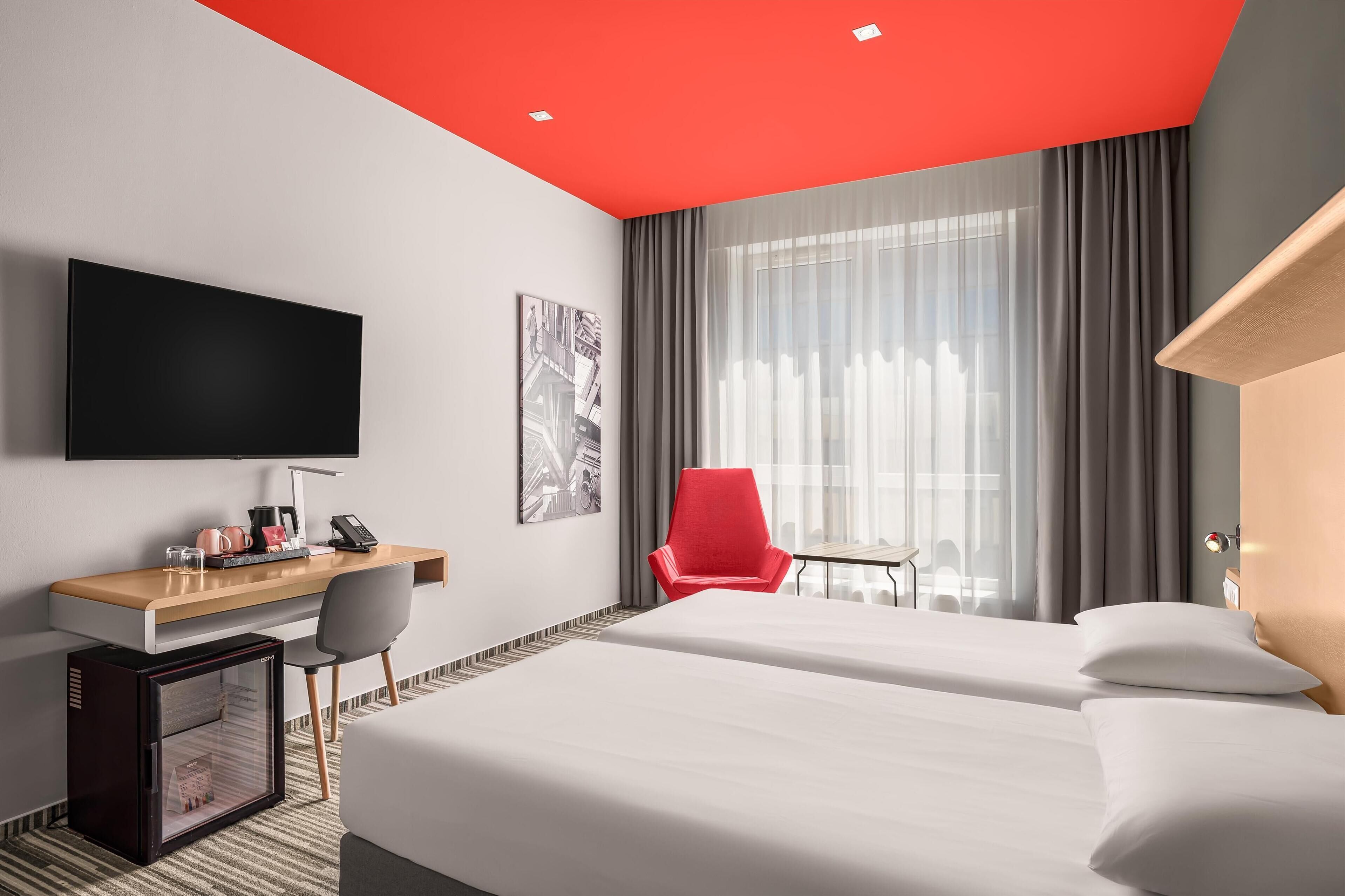 Hotellihuone, jossa on sänky, työpöytä tietokoneella, televisio ja punainen tuoli.
