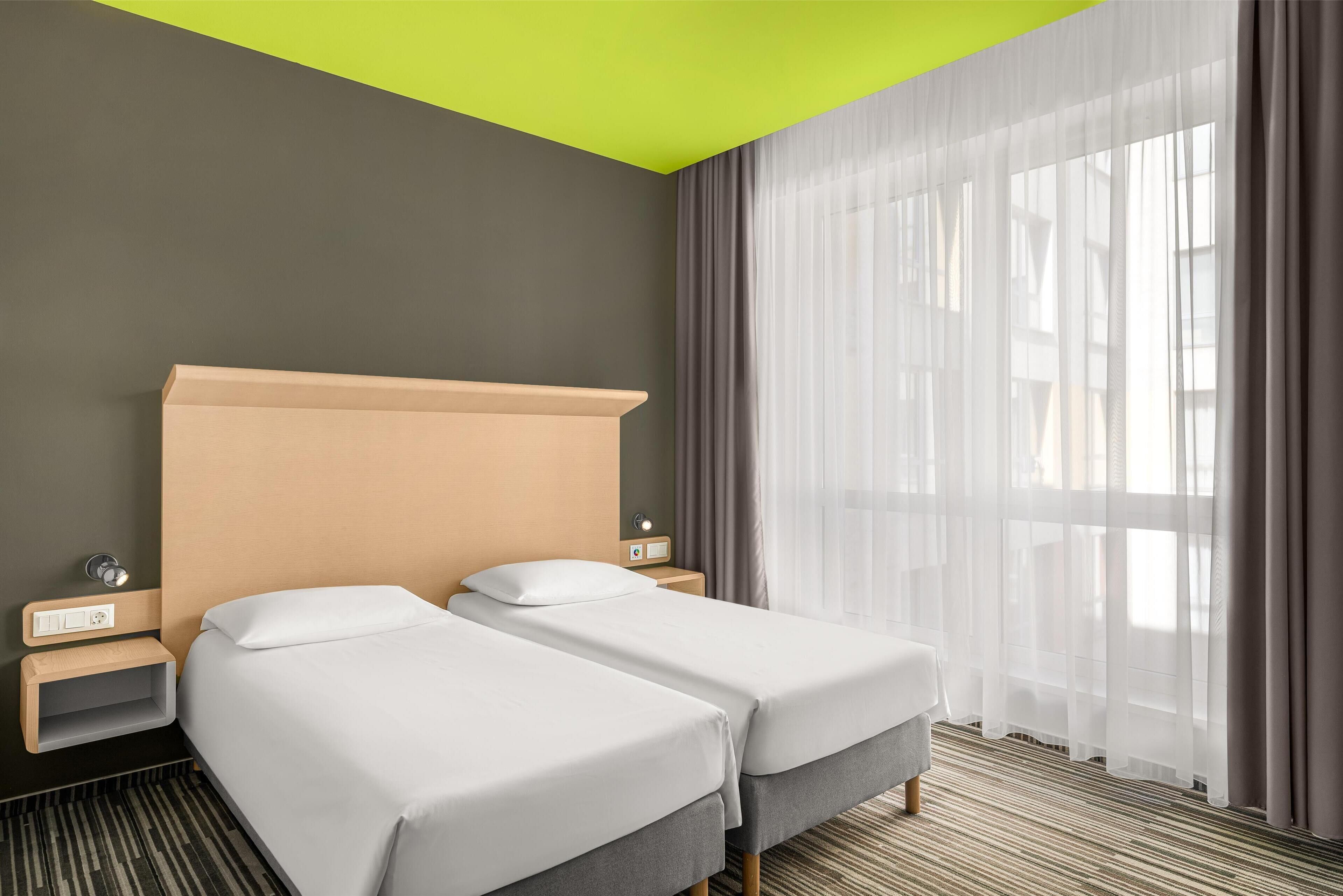 Een hotelkamer met twee bedden, een heldergroen plafond, een groot raam met dunne gordijnen en een gestreept tapijt.
