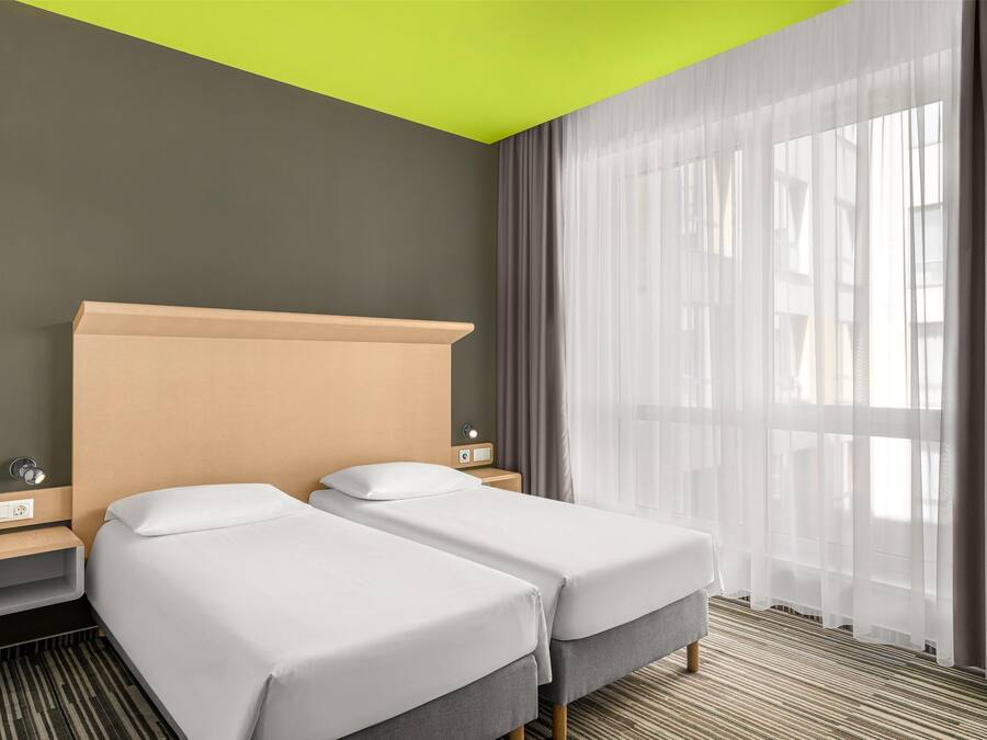 Een hotelkamer met twee bedden, een heldergroen plafond, een groot raam met dunne gordijnen en een gestreept tapijt.