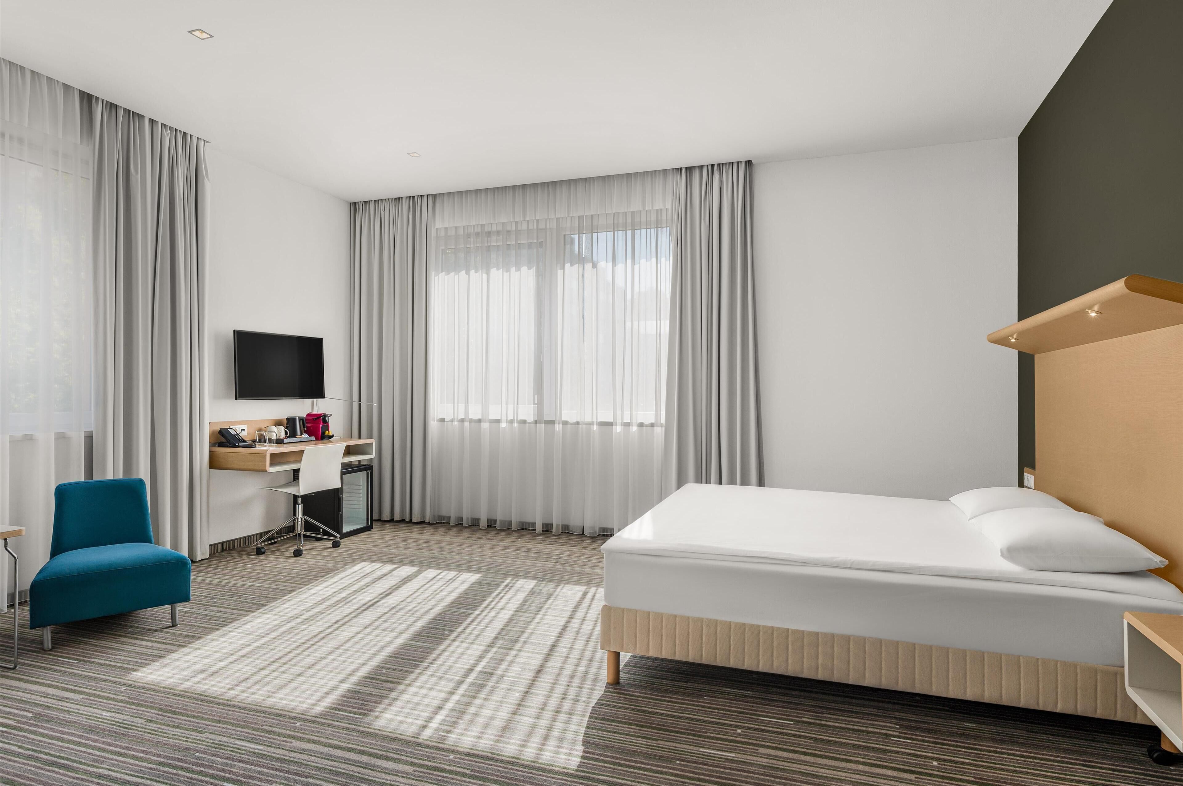 Habitación de hotel con cama, escritorio con computadora, silla, televisor y ventana con cortinas.