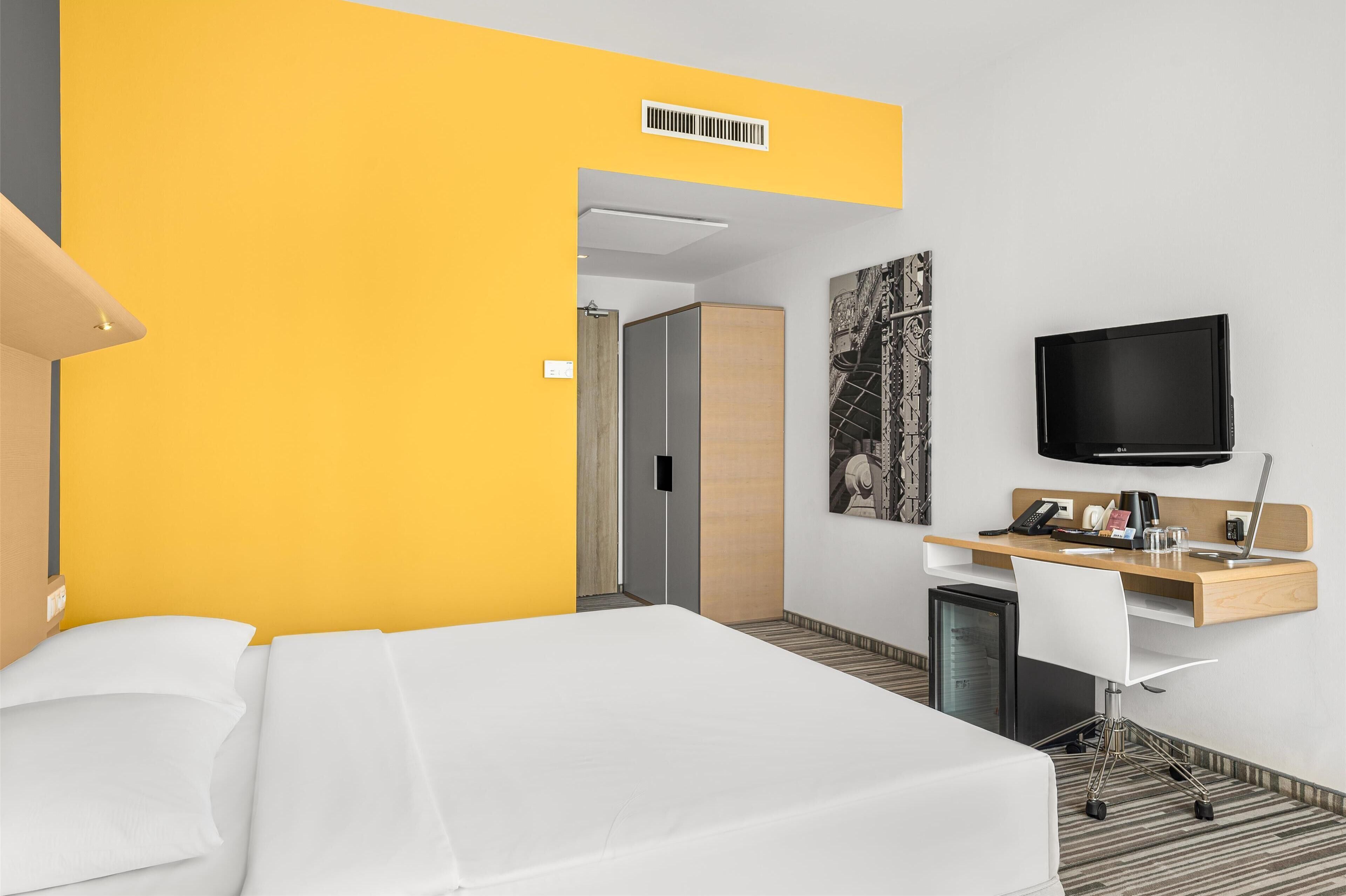 Une chambre d’hôtel équipée d’un lit, d’un bureau avec un ordinateur, d’une télévision et d’un petit réfrigérateur.
