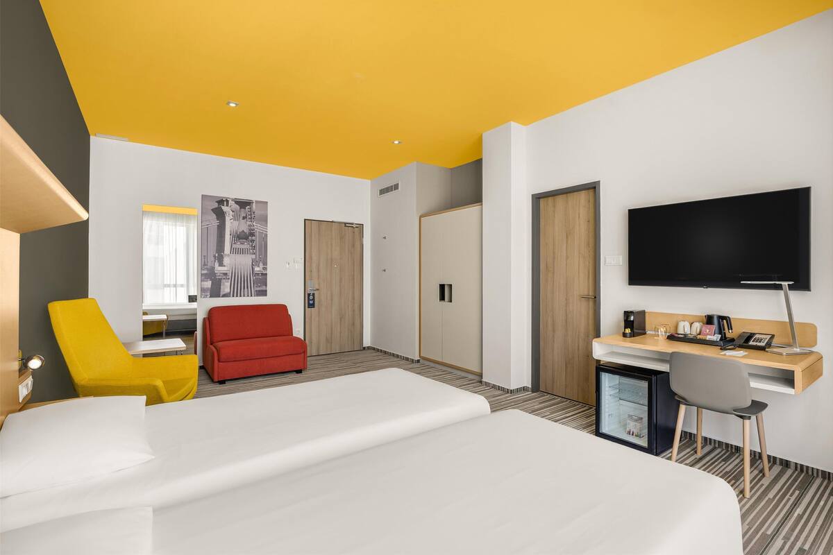 A modern hotel room with a bed, a desk, a TV, a mini-fridge, and a red chair.