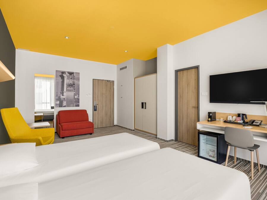 A modern hotel room with a bed, a desk, a TV, a mini-fridge, and a red chair.
