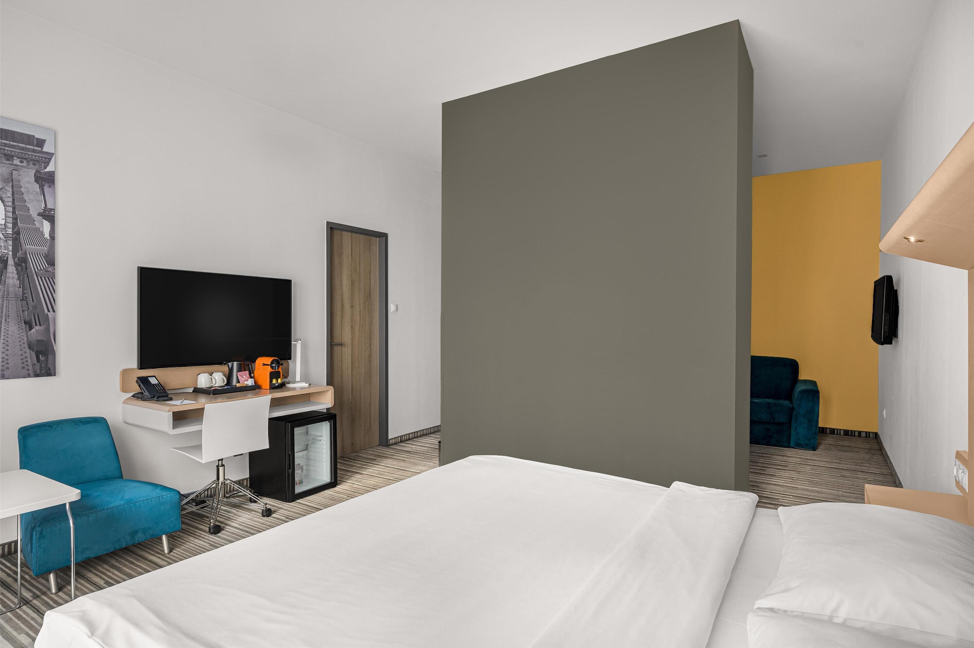 Una habitación de hotel moderna con una cama grande, un escritorio con televisor de pantalla plana, una silla azul y una kitchenette pequeña.