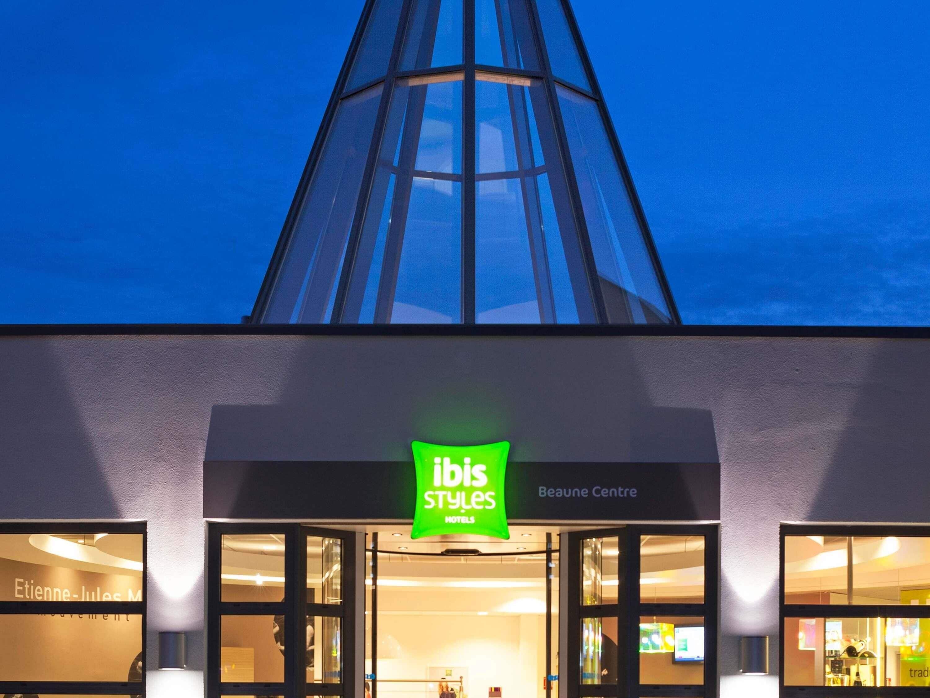 A hotel entrance with a modern glass facade, a green sign for 'ibis Styles Hotel,' and a 'Beaune Centre' sign above.