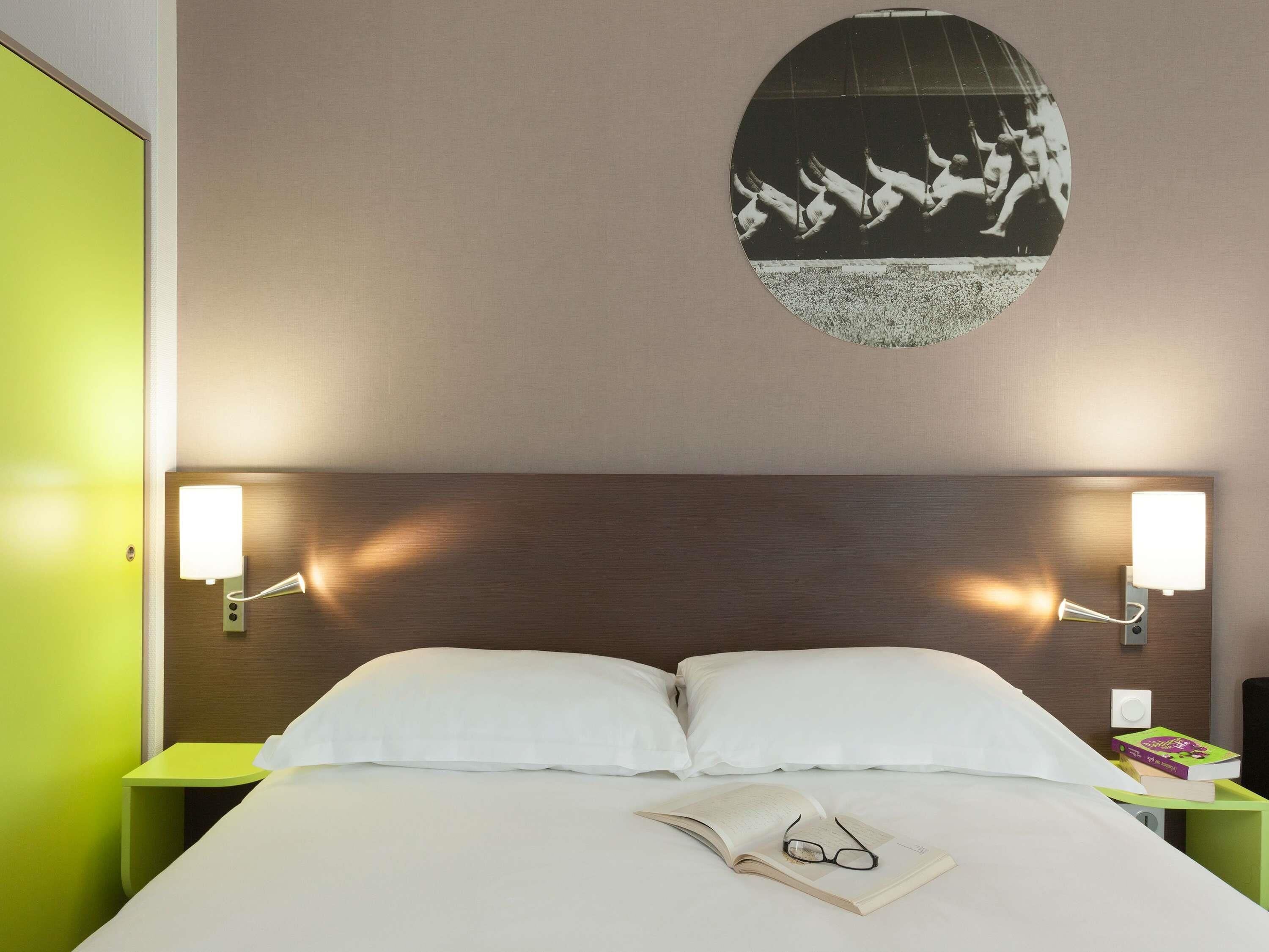 A hotel room with a bed, two bedside lamps, a wall-mounted light fixture, and a wall art piece.