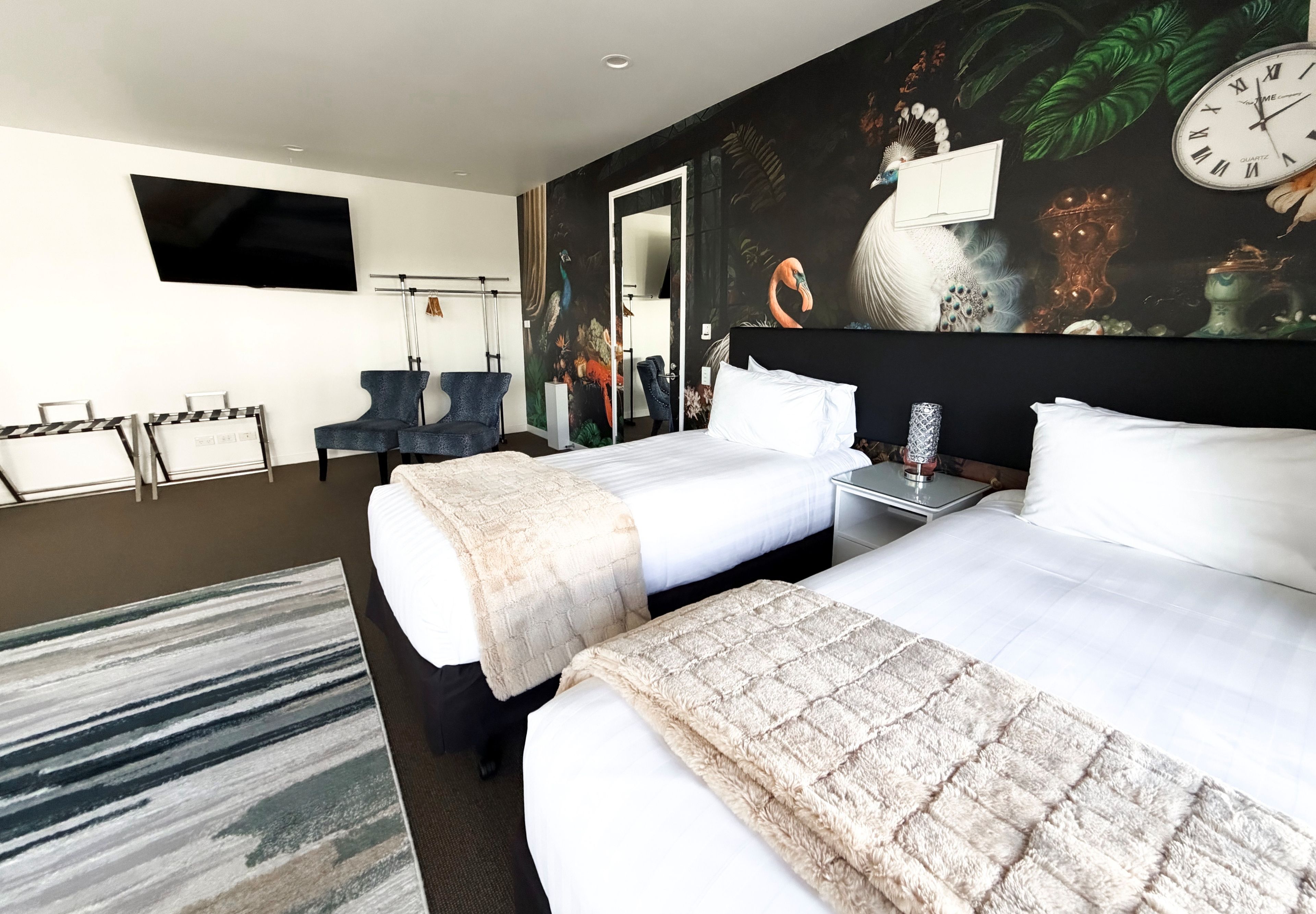 Ett hotellrum med två sängar, en platt-TV, en tavla och en väckarklocka på väggen.