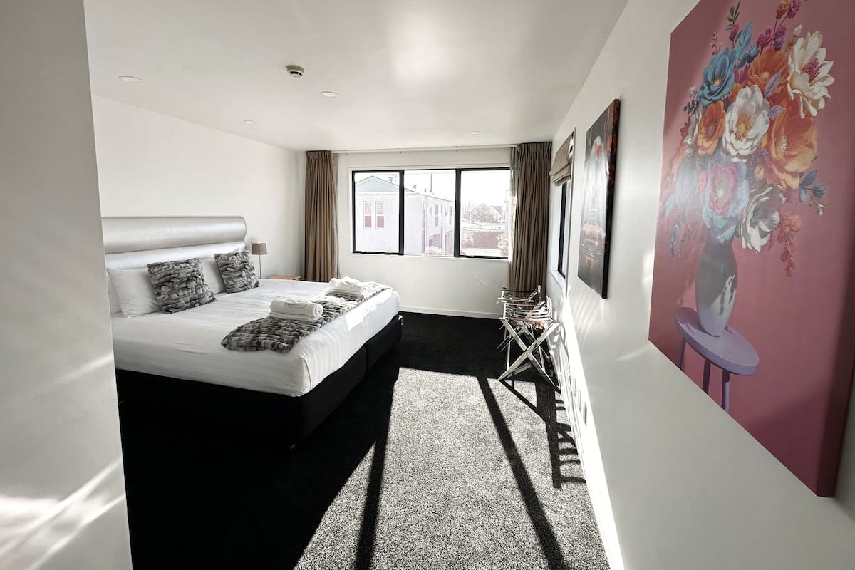 A modern hotel room with a large bed, a window with curtains, a painting on the wall, and a small table with chairs.