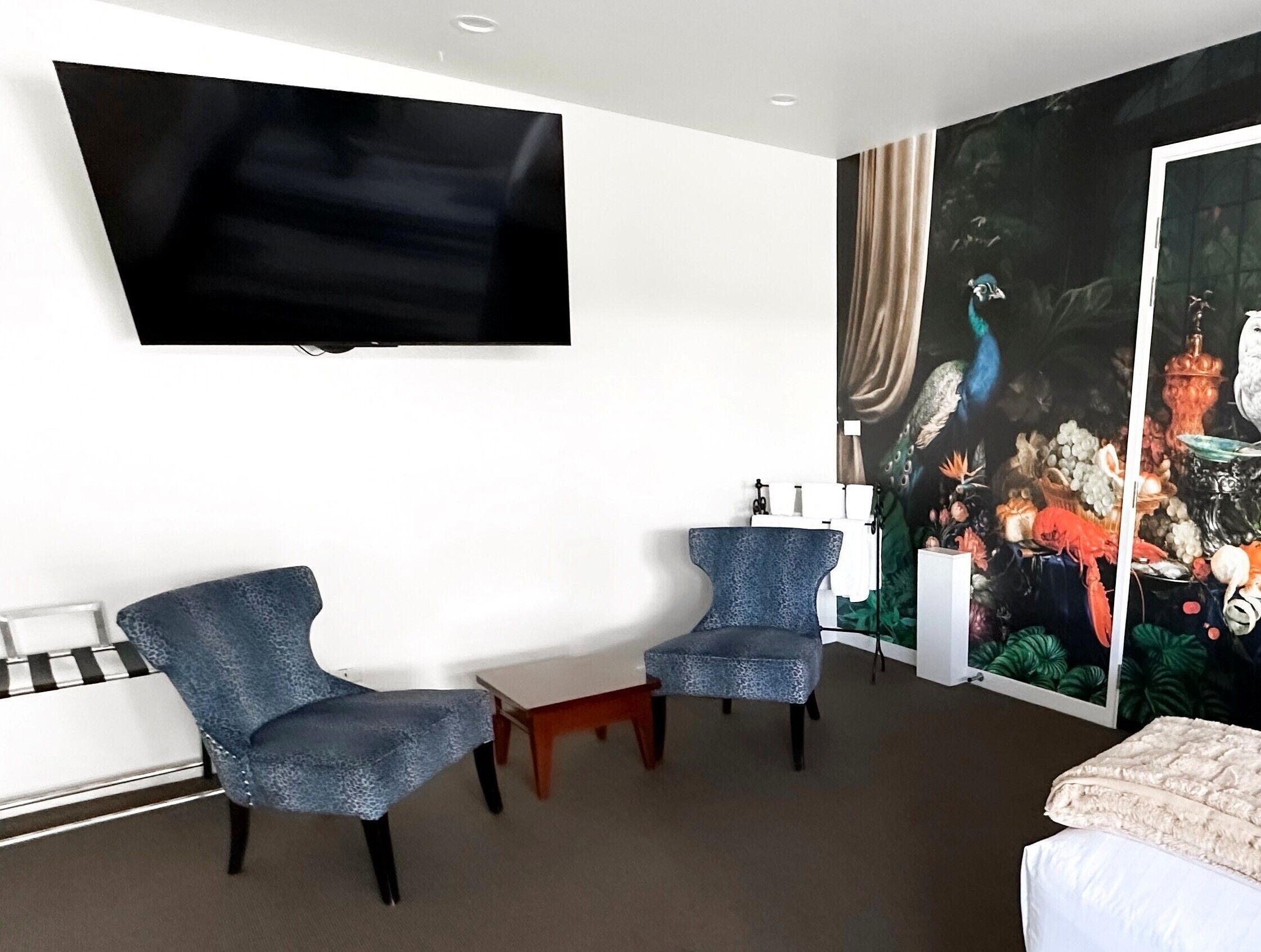 A hotel room with a large flat-screen TV mounted on the wall, two blue armchairs, a white bed, and a vibrant mural on the wall.