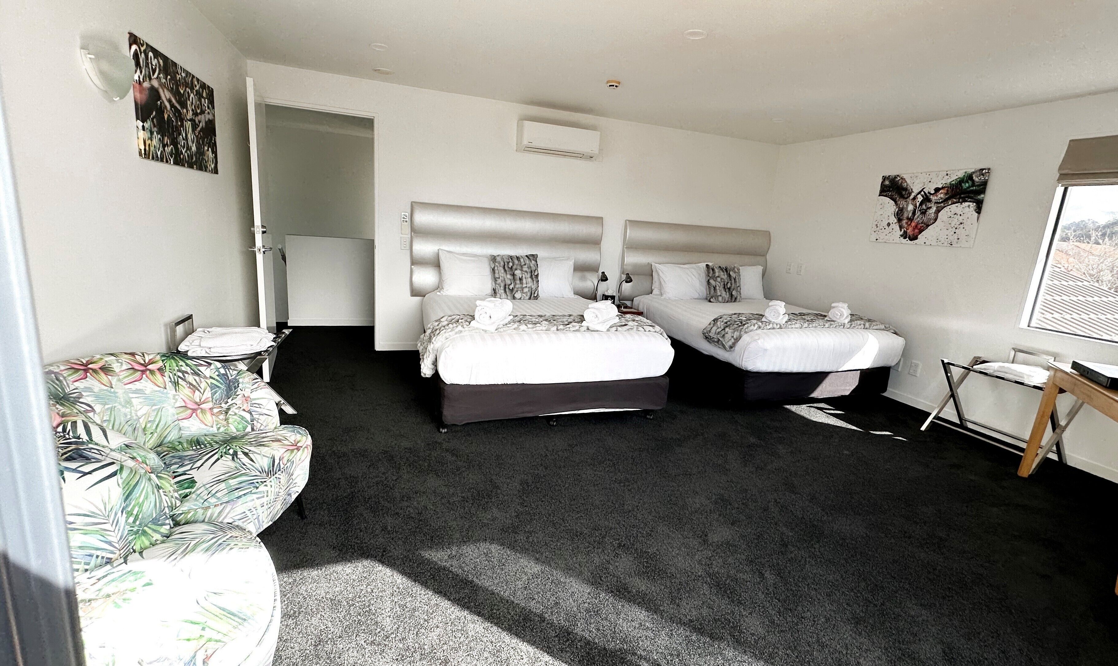 A hotel room with two beds, a small table, a chair, a window, and artwork on the walls.