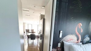 A hotel room with a bed, a painting of a flamingo, and a view into the dining area. - DeLorenzo's Studio Apartments (Nelson)