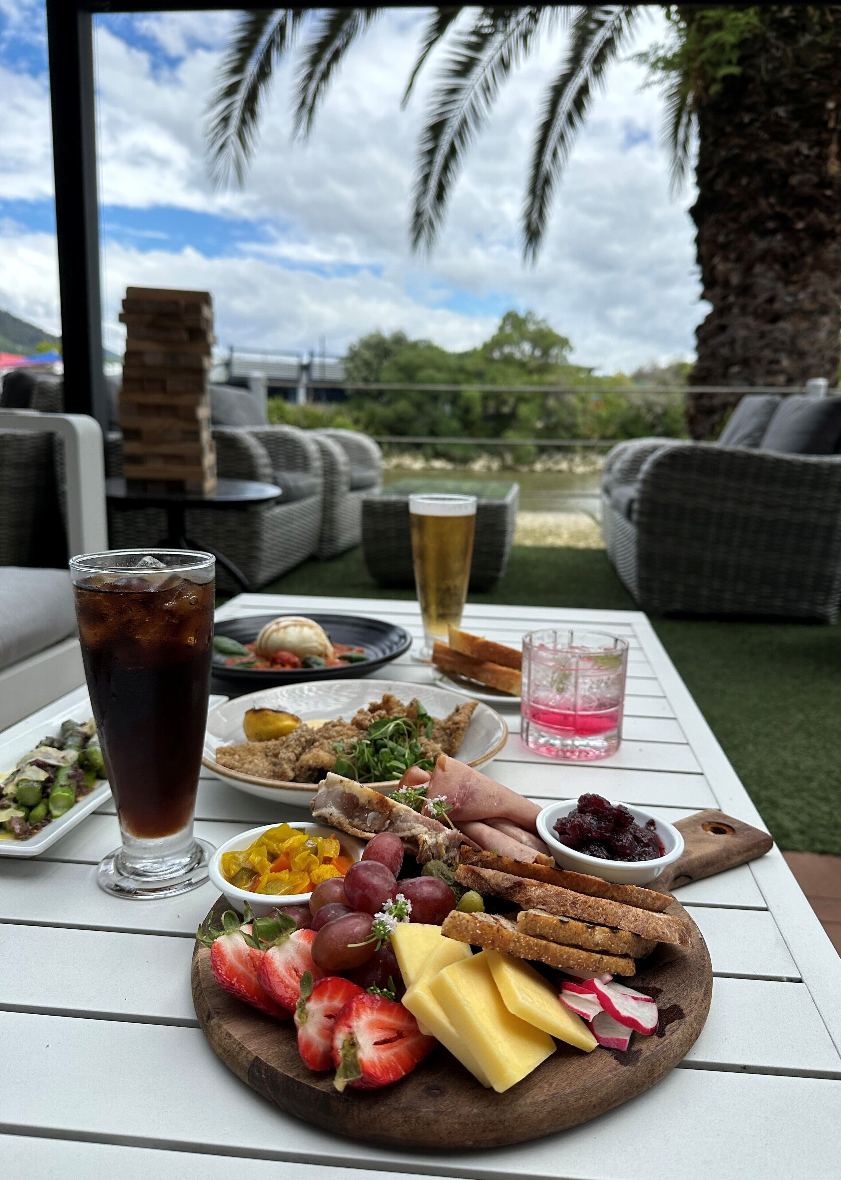 A table set with a variety of foods and drinks, including a glass of beer, a glass of iced tea, and a platter of fruits and cheeses, with a view of palm trees and greenery in the background.