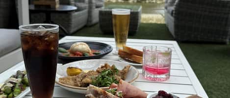 Una mesa servida con una variedad de comidas y bebidas, incluyendo una cerveza en vaso, un té helado y una bandeja de frutas y quesos, con vistas a palmeras y follaje al fondo.