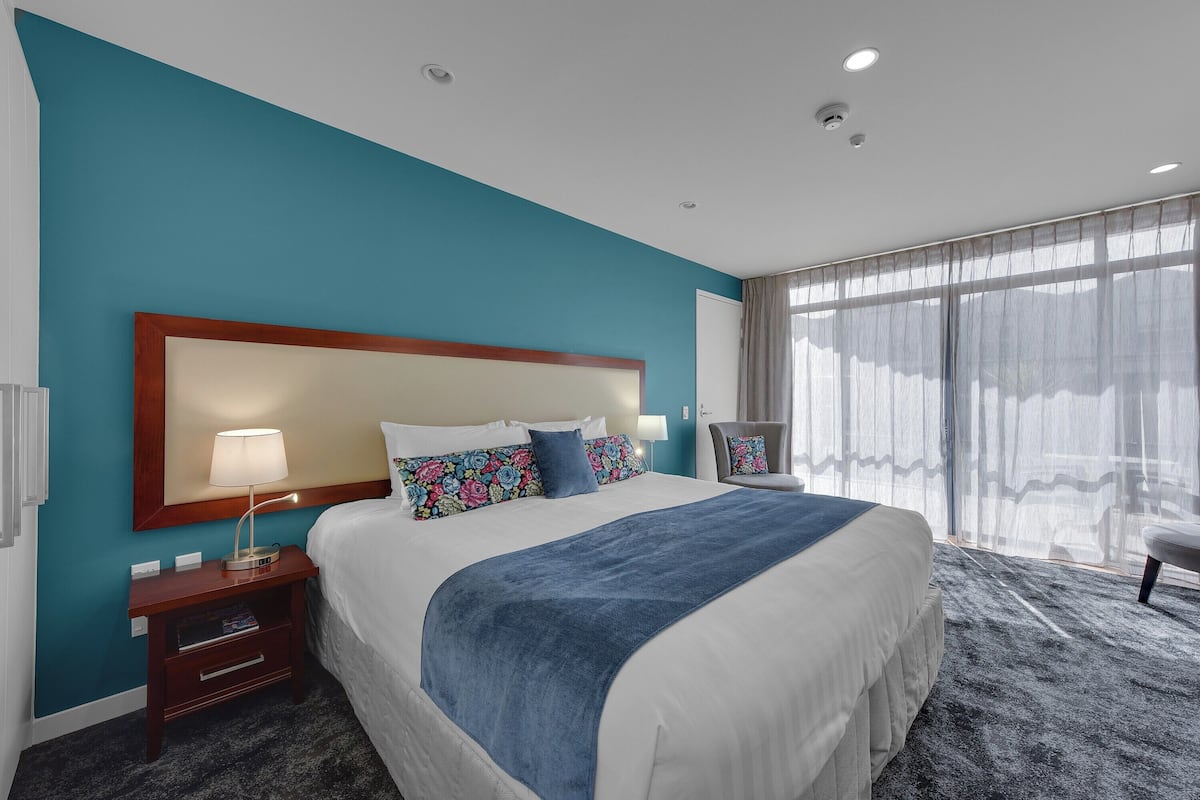 A hotel room with a large bed, a nightstand with a lamp, a chair, and a large window with curtains.