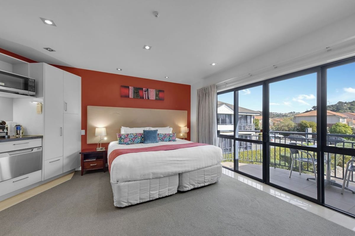 A modern hotel room with a large bed, a kitchenette, and a balcony with a view of the city.