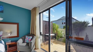 A hotel room with a bed, a desk, a chair, a sofa, and a sliding glass door leading to an outdoor patio with a table and chairs. - DeLorenzo's Studio Apartments (Nelson)
